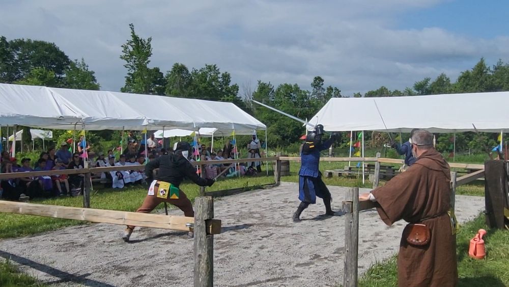 The woman in the blue outfit is now wearing a helmet. She is between two swordsmen, defending herself with her greatsword, which os raised above her head. An audience is watching  the demonstration