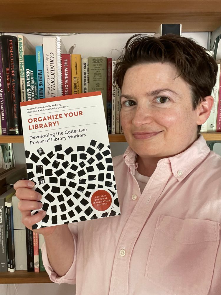 Me in a pink button-up shirt holding a copy of our book, “Organize Your Library! Developing the Collective Power of Library Workers.”