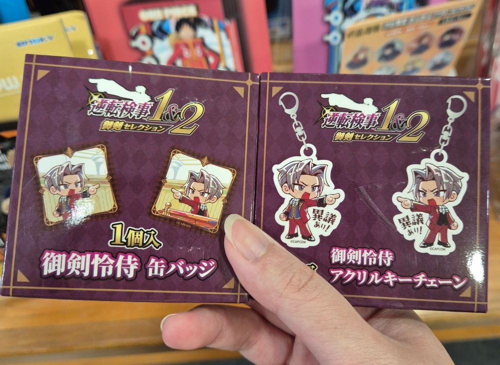 A photo of 2 blind boxes containing Miles Edgeworth merchandise, the left contains a badge and the right has an acrylic keychain.