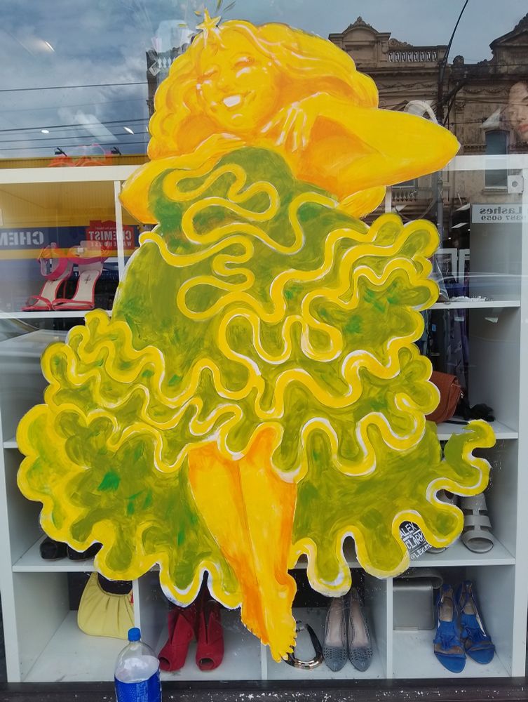 Big window painting of a bright yellow lady wearing a green dress with yellow tinsel wiggly lines all over it