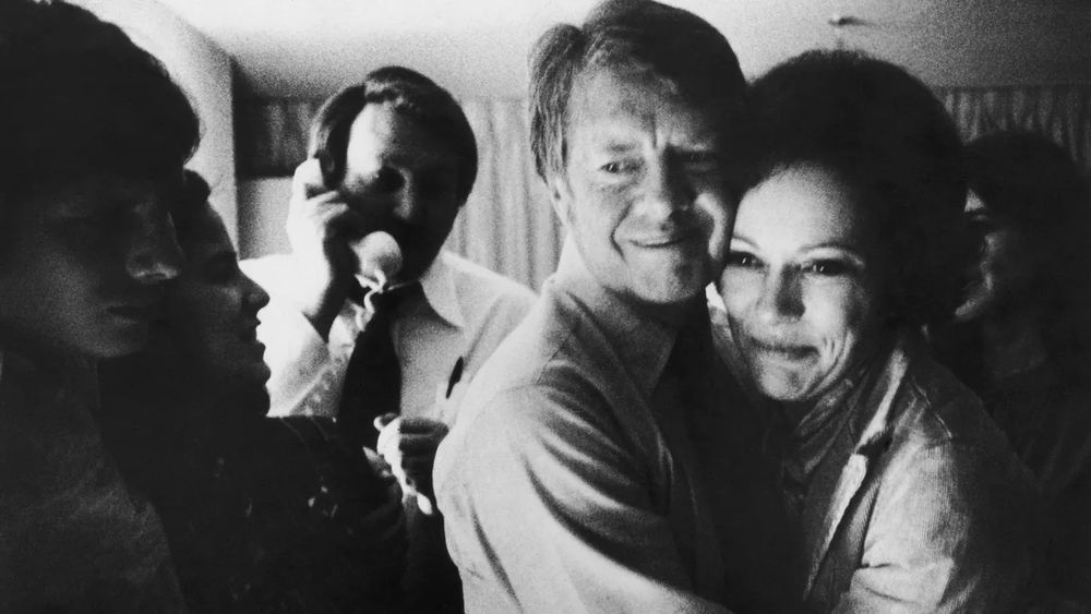 A black & white photograph of Jimmy and Rosalynn Carter embracing in private with their family after winning the 1976 presidential election. Their smiles are warm yet restrained. There is a campaign staffer in the background on the telephone. The lighting illuminates only on Jimmy and Rosalyn’s faces clearly yet softly, everyone else’s faces are in shadow. It feels warm and safe