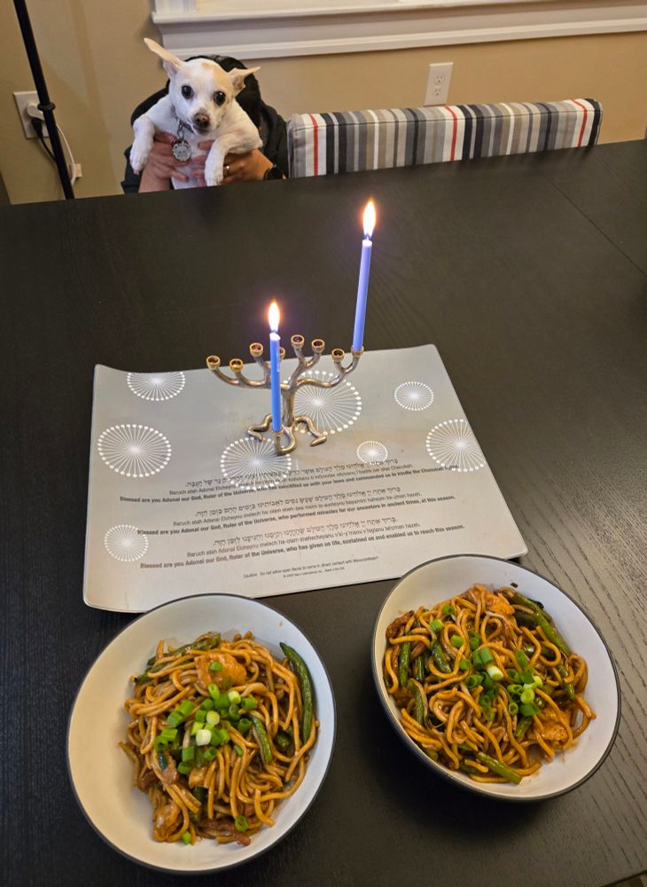 A menorah lit for the first night of #Hanukkah is on a black table. In front of it are two bowls of Chinese food. Behind the table, Sebastian the dog is held up so you can't see who's behind him. 