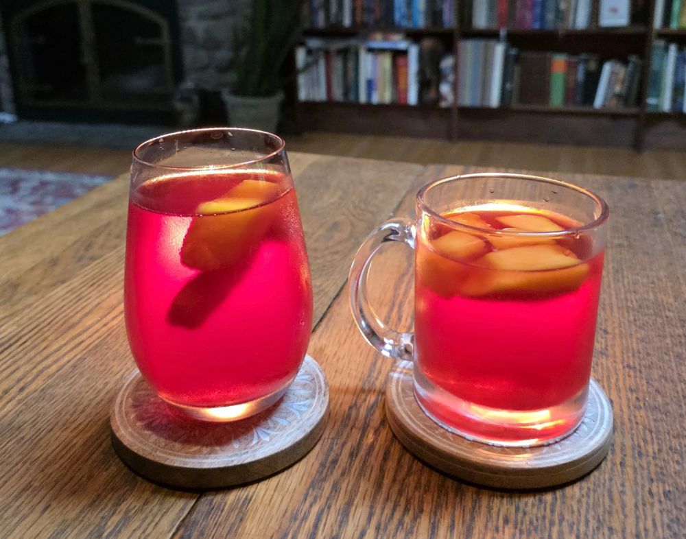 Served cold! Two vibrant pinkish-red cups of hibiscus tea in clear glasses - cold, not hot, with frozen mango chunks floating on top. Backlit by tealights (hidden behind the mugs), sitting on a wooden table with a bookcase in the background.