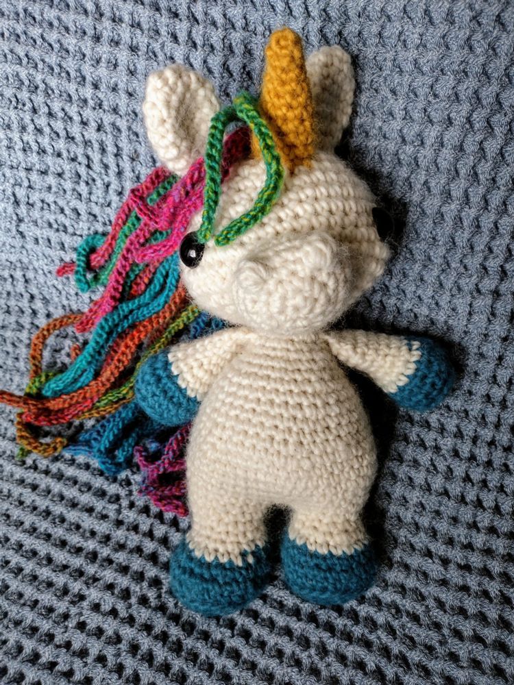 a white crochet unicorn with a yellow horn, blue hooves, and a rainbow mane, resting against a light blue waffle-textured blanket