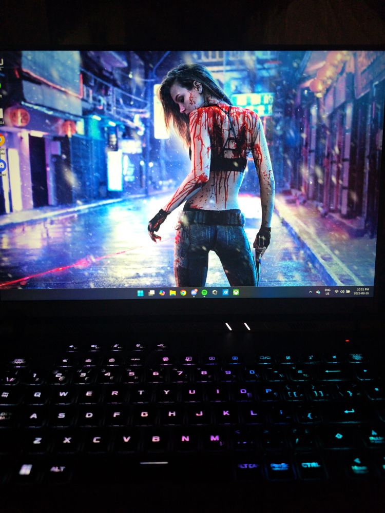 Trans coloured backlit keyboard and a desktop wallpaper depicting a dark haired girl in a halter top, covered in blood (it doesn't appear to be hers), holding a pistol in her right hand. The perspective is from the back and she is looking at a laser beam that is pointing at her left thigh.