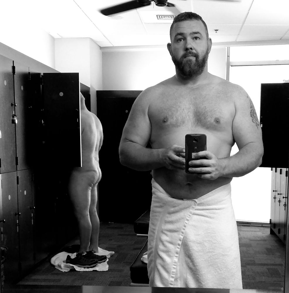 Photo is black and white. Man with a towel wrapped around his waist in the foreground of a gym locker room. Another man is naked behind him to his right getting his clothes from a locker.