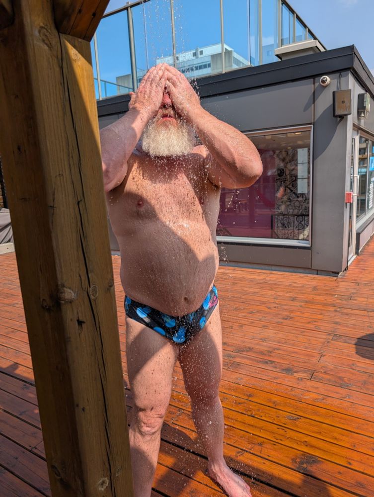 A man rinsing off in an outside shower in the mid day sun. He's wearing a swim brief with blue jellyfish on it.