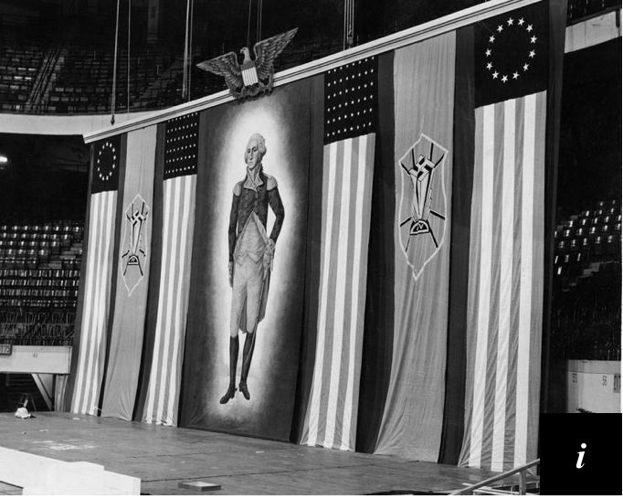 A black and white picture of the backdrop of the February 1939 German American Bund fascist rally at Madison Square Garden. At center, an idealized Washington in military uniform floats standing in a glow on a darker background. The portrait is flanked on either side by two American flag banners with then current and 1776 style star fields. Between each pair hang banners with the Bund’s Nazi swastika topped icon on a shield on a red background. Above the Washington portrait is an American eagle with spread wings stylized in the manner of the Nazi eagle.