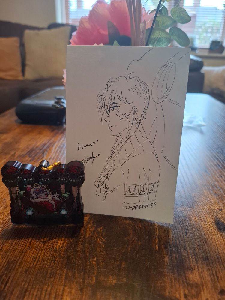 Photo of the charm next to a drawing of Icarus from Hades II 