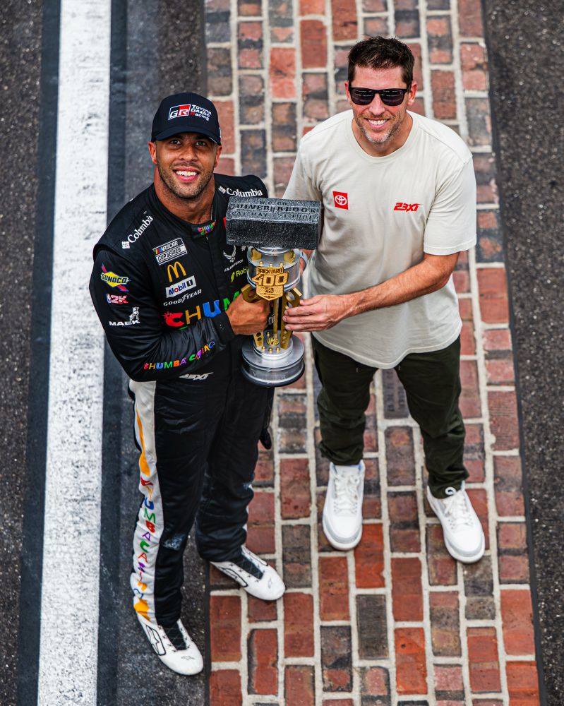 Bubba Wallace and car owner Denny Hamlin pose on the yard of bricks, smiling up towards the flag stand with the race trophy.