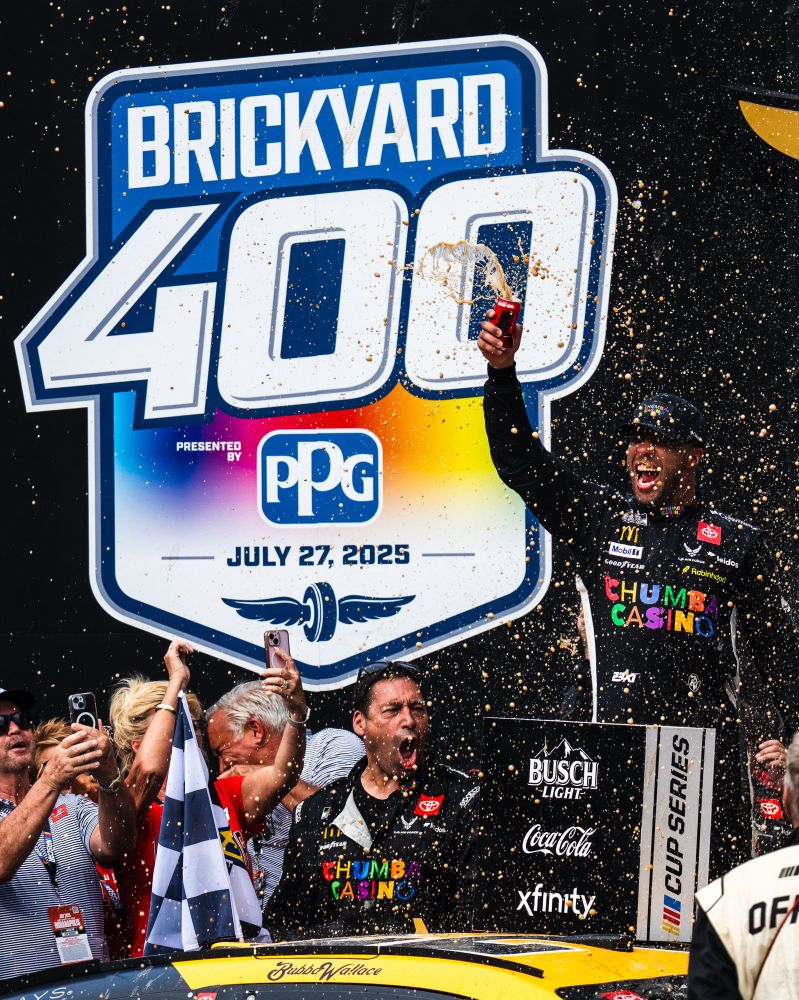 Bubba Wallace splashes soda all over as he celebrates in victory lane in front of vibrant Brickyard 400 signage