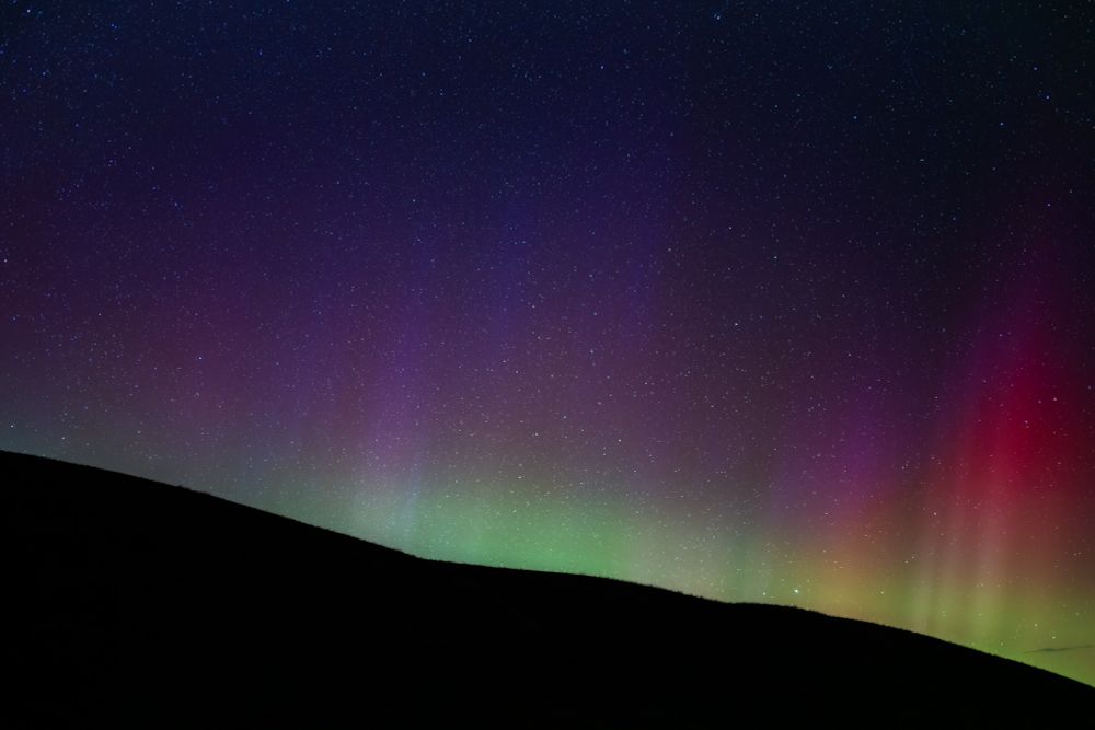 Vibrant aurora colors glimmer near the horizon. A foreground hill leaves a diagonal shadow at the bottom of the frame.