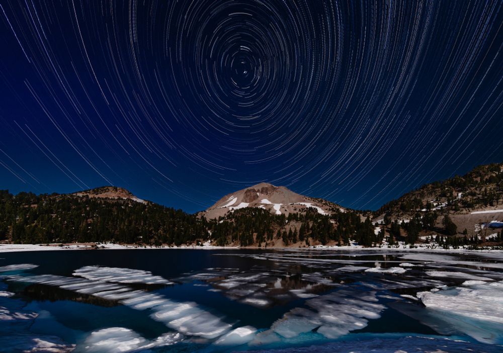Star trails swirl over a horizon defined by Mount Lassen as winter remnant ice chunks float in a foreground alpine lake.