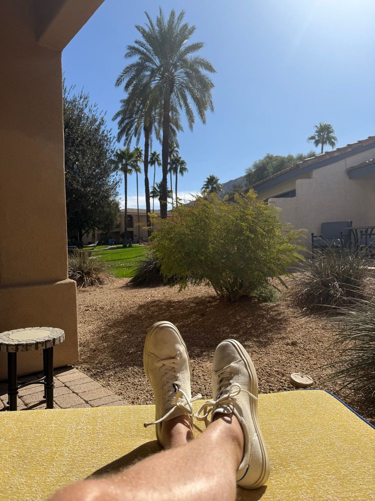 A relaxed scene showing a person’s legs stretched out on a lounge chair with white sneakers. Palm trees, green shrubs, and a desert-style landscape surround a building with a terracotta-tiled roof in the background.