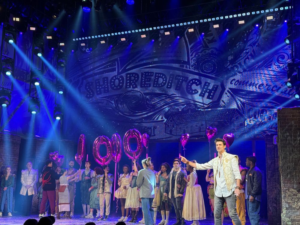 A vibrant stage scene from the musical & Juliet, featuring the cast celebrating a milestone performance. The backdrop reads “Shoreditch” with vivid lighting effects and street-style art. Cast members hold shiny pink balloon letters spelling “1000!” and heart-shaped balloons. Performers are dressed in a mix of modern and period costumes, with joyful expressions.