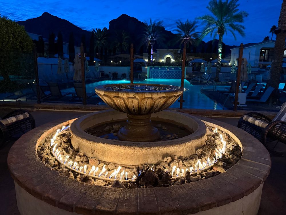 A lit fire feature surrounds a stone fountain in an outdoor lounge area at night. In the background, a swimming pool, palm trees, and a silhouette of mountains are visible under a deep blue sky.