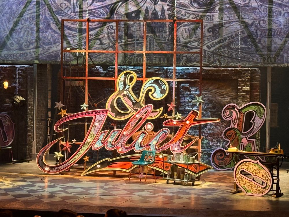 A detailed stage set for & Juliet featuring a large, colorful neon-style sign that reads “& Juliet” in ornate lettering with stars and flourishes. The backdrop includes brick walls and a vintage London street map. The floor is checkered, and set pieces include paint cans and scaffolding, creating a whimsical, creative atmosphere.