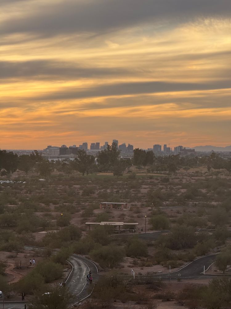 A scenic view from Hole in the Rock, Papago Park, showing a winding desert trail, picnic shelters, and Phoenix’s downtown skyline in the distance under a golden sunset sky.
