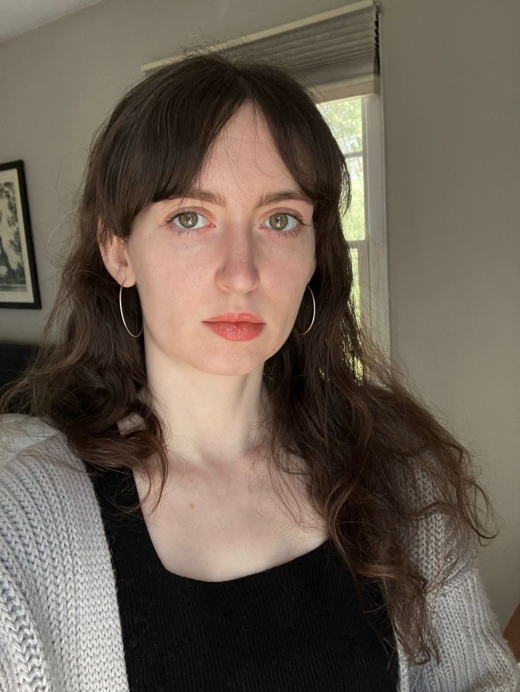 a selfie. Amelia is a white person in her twenties with brown hair and parted bangs, wearing a black square neck top, a gray cardigan, and silver hoop earrings