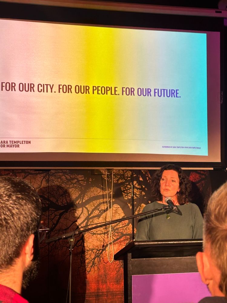 Mayoral candidate Sara Templeton delivering a speech at her launch on Friday. Sara has curly dark hair and is wearing a blue top, standing at a podium with the edge of her purple campaign poster just visible. Behind her is a screen with her "For Our CIty. Four Our People. For Our Future" statement displayed in the center of it. There's a projected outlines of a leafless tree below the screen. The backs of the heads of two audience members are visible on the left and right of the picture. 