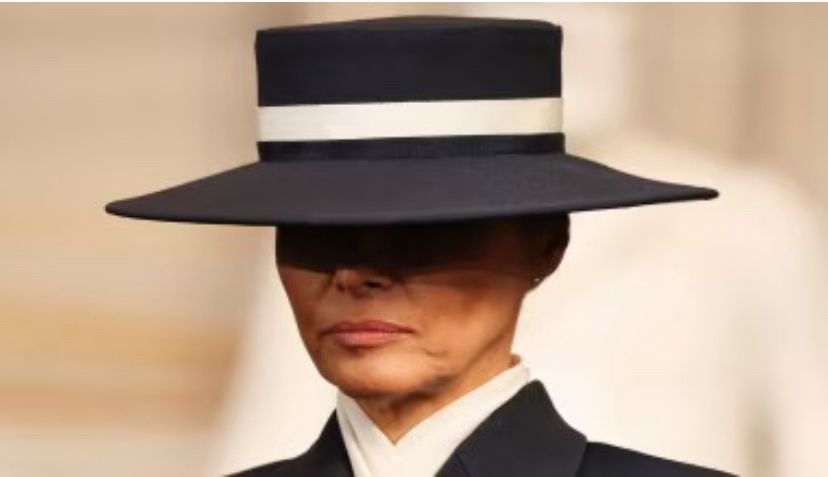 Melania wife of Cheeto face wearing dark wide brim hat and covering to the neck 