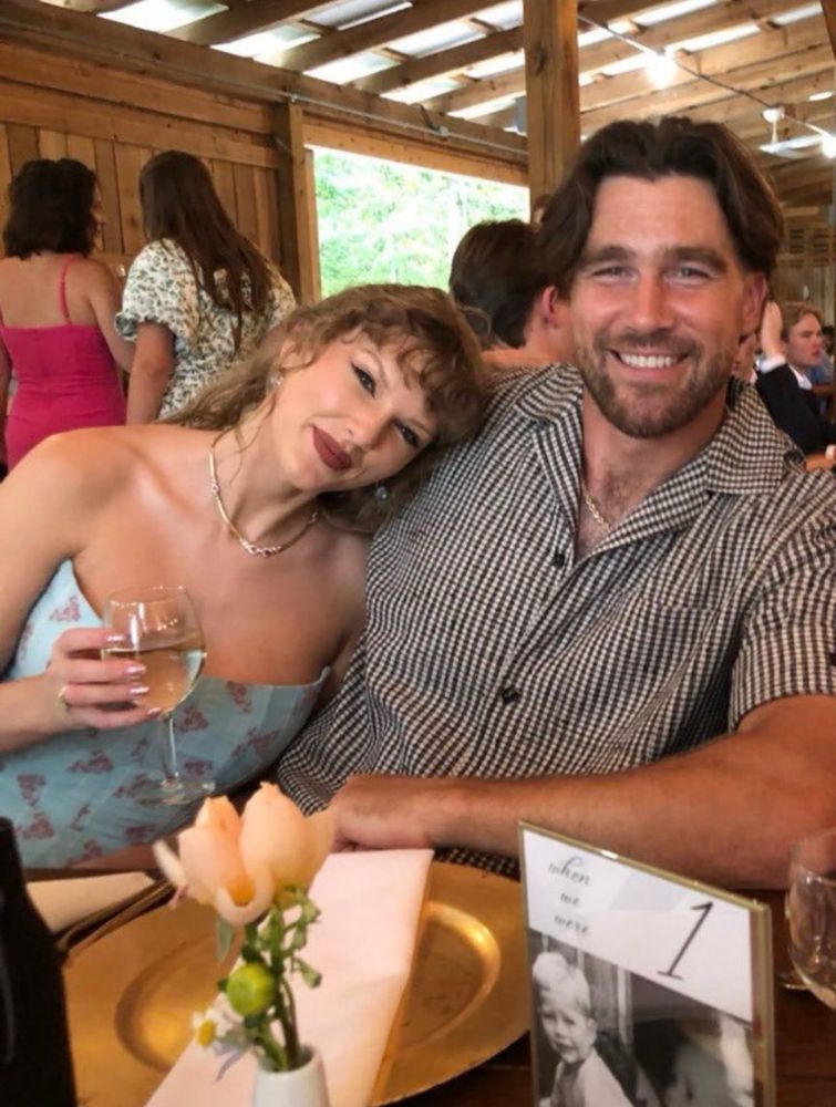 Photo of Taylor Swift and Travis Kelce. Taylor is wearing strapless blue dress holding a glass of wine in her hand. Travis is wearing a black and white patterned buttoned up shirt. They have there arms around each other  