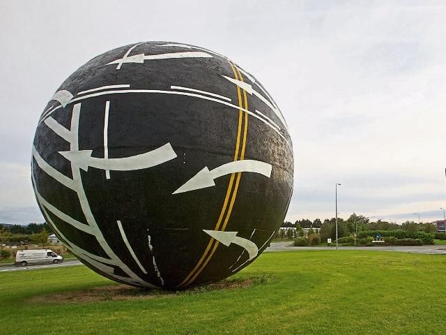 a giant ball, about the height of a house, sits in a grassy roundabout. It's a black ball which appears to be made of asphalt and includes road markings such as double yellow lines, continuous white lines, broken white lines, and arrows. It looks like maybe a giant scraped up the road and rolled it into a smooth ball between their fingers and left it there