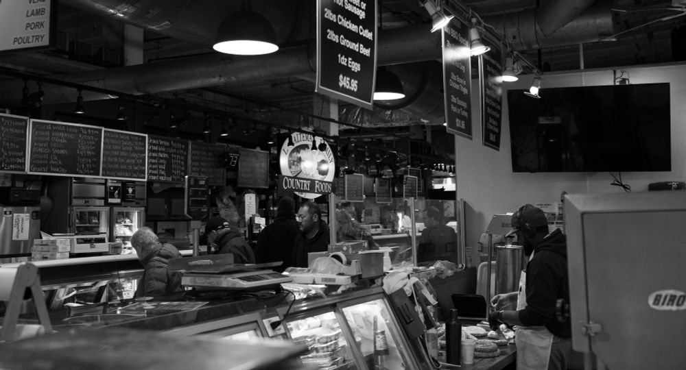 A black and white photo at meat counter level in a busy market (Reading Terminal), a man fills sausage casings while other people pursue an adjacent display case. 