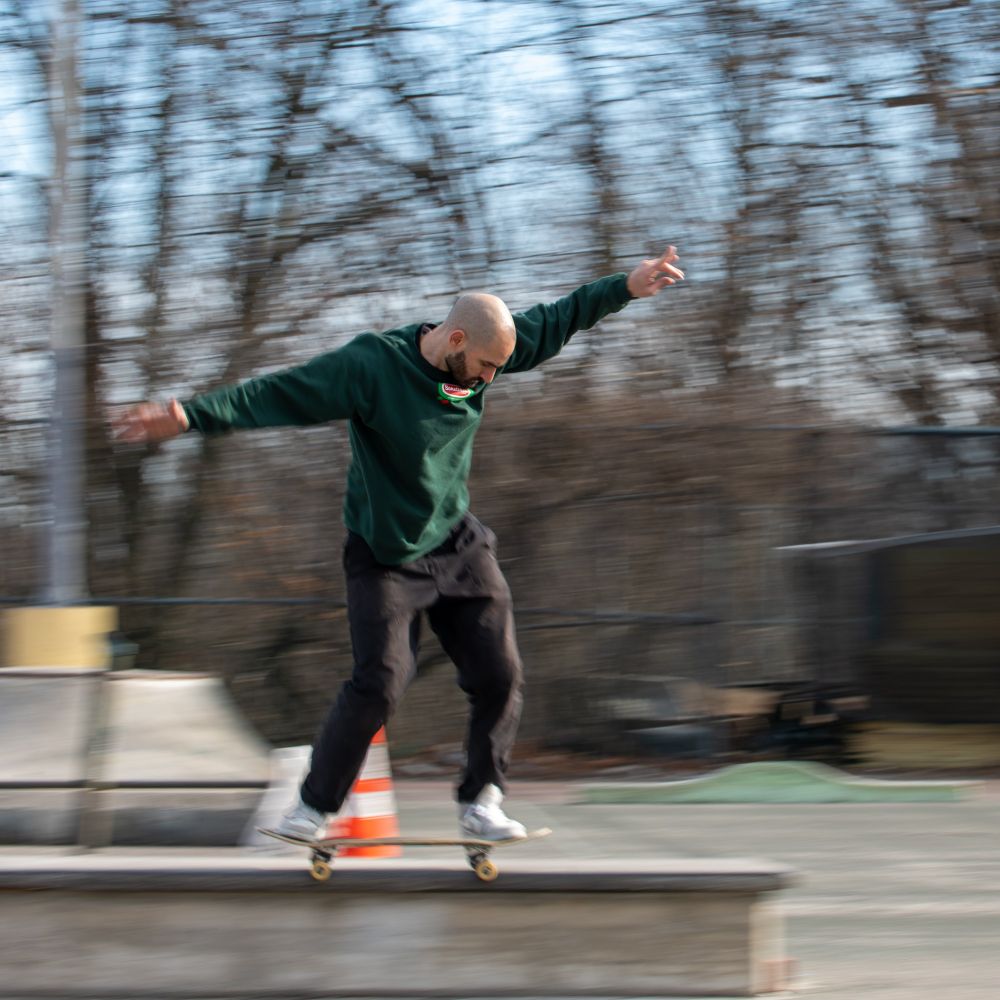 Photo of a bald man in a green crew neck doing a 50-50 backside grind. All except for the man's upper torso is blurred due to the motion of the camera movement.