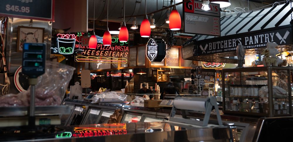 Another photo at meat counter level, this one in color, a man in the center of the frame prepares a cheesesteak far across the market, in the mid ground a woman places her order at the meat counter, and in the foreground sausages are being weighed.