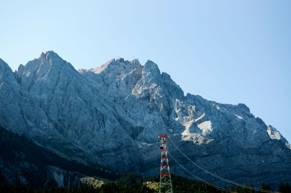 The Zugspitze mountain with a cable car tower in the foreground. A cable car is on its way up and you can barely see a crane at the peak.