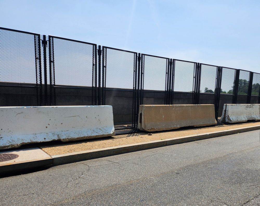 Black vertical fencing 8ft tall lines a small bridge along the Tidal Basin.  There are several stand-alone jersey barriers in front of the fences.
