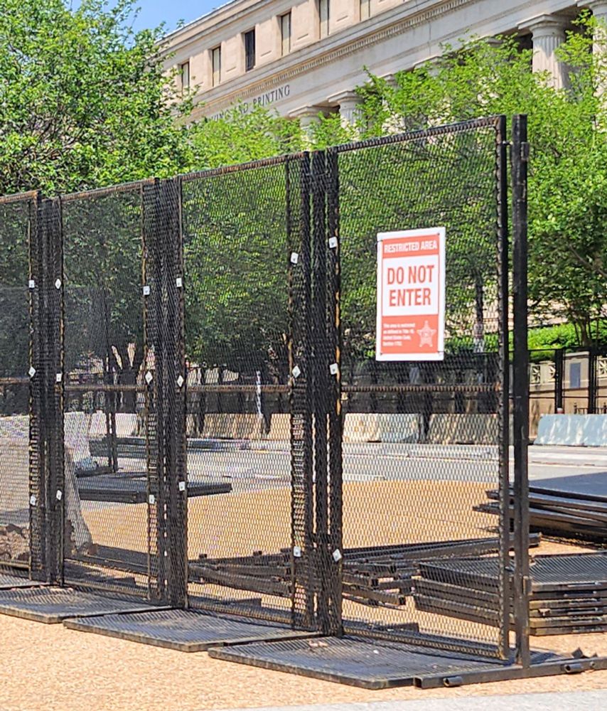 Vertical black fencing, roughly 8 ft tall, with a sign saying "Restricted Area. Do Not Enter." Each section of fencing also has a counterweight that extends about 2 feet away from the fence in both directions. 

There is a gap in the fencing, where more stacks of fencing are laid out to be erected.