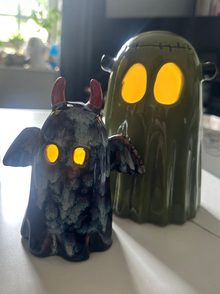 two ceramic ghosts, one slightly smaller than the other, with light up eyes. the smaller one is black with grey mottled paint on it and devil horns and wings, while the larger one is modelled after frankenstein's monster - it is green with stitches along its head and two bolts on either side of the head 