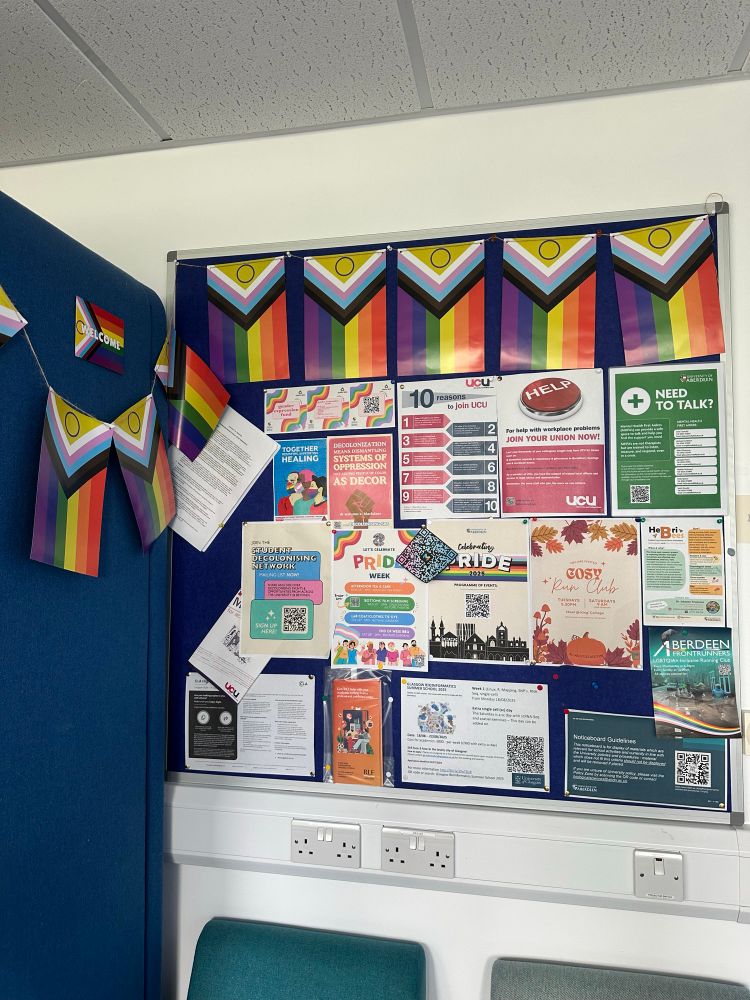 blue noticeboard covered in various posters and flyers and decorated with progress flag bunting along the top and side. many of the posters are related to pride month, as well as others advertising various activities around the department/uni/local community
