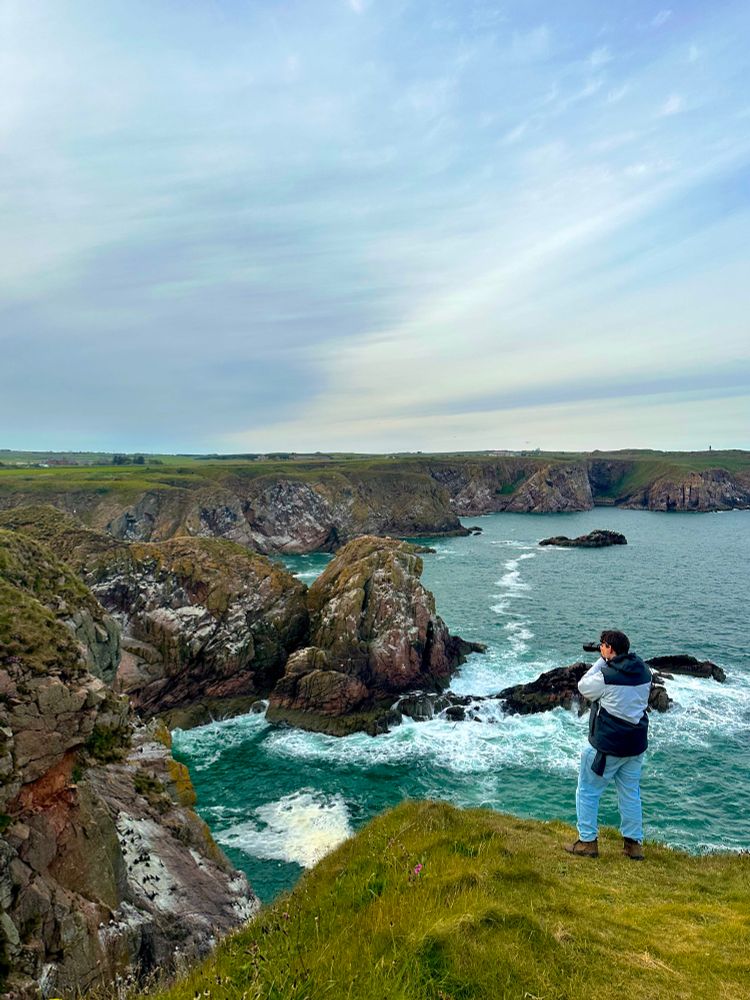 multiple jagged cliff edges overlooking clear blue ocean waves. a person is stood on the nearest cliff edge photographing the sea birds that fly along the cliffs below us 