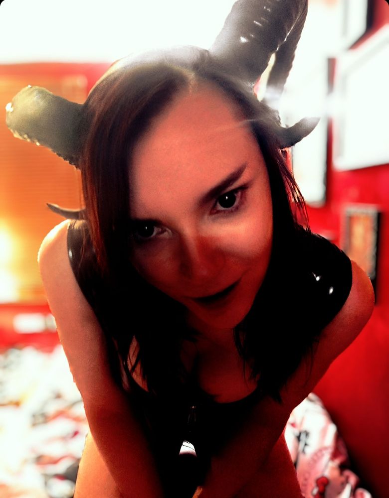 A boy or girl demon with hornw looking intently into the camera wearing a shiny black dress but with his or her arms out. High conrast. Much horn.