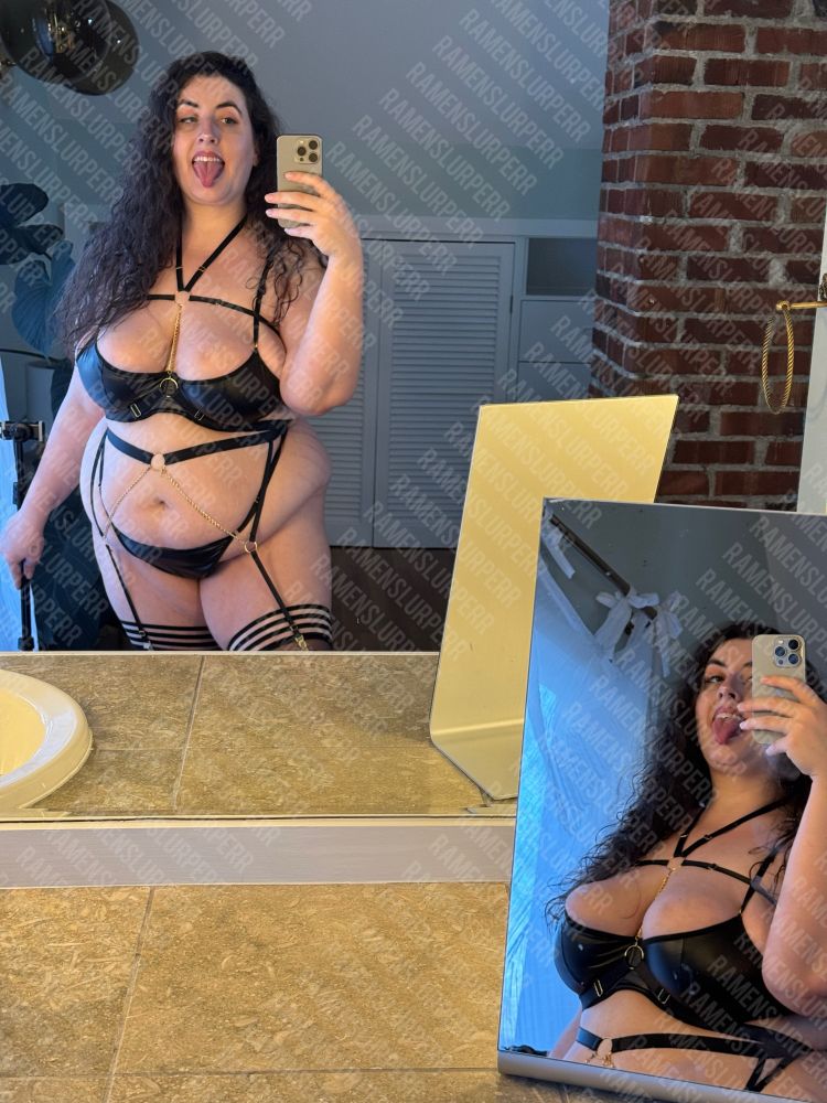 BBW Marie stands in front of a large mirror with another mirror on a countertop reflecting her image back two times. She wears a black strap lingerie set with gold chain down the belly and chest and black thigh high stockings. her fat stomach spills out over the top her large breasts push against the fabric of her bra. Her hair is loose and long. She sticks out her tongue. 