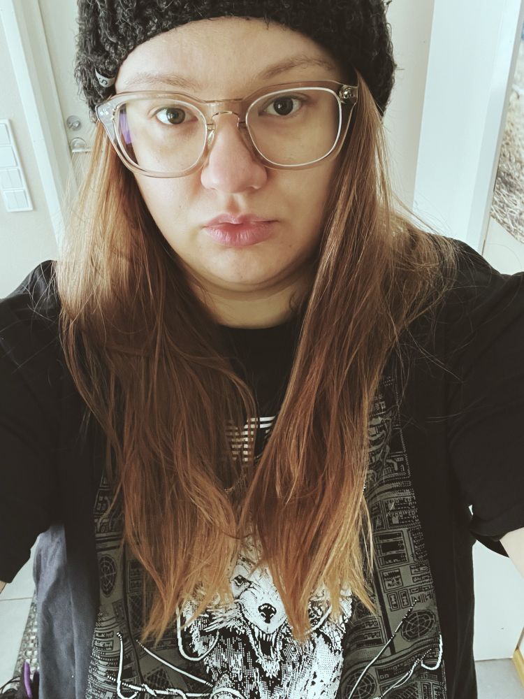 Woman with glasses, brown eyes, light brownish hair and light skin dressed in a black band t shirt and a beanie. 