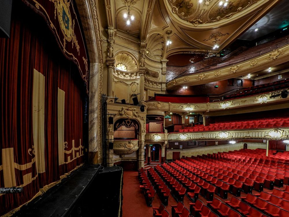 Side view of historic theatre auditorium, showing part of the stage with a dark red curtain, rows of bright red seats in the stalls and dress circle and balconies and box with cream and gold plasterwork.
