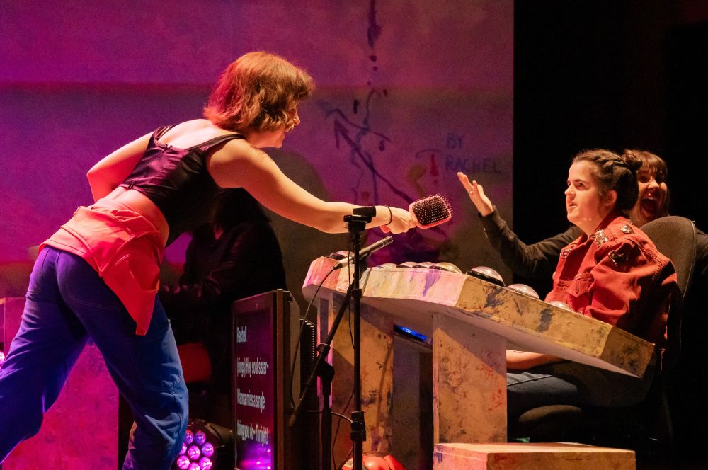 Flo leans across a colourful desk with a hairbrush in her hand like a microphone. Sat behind the desk is Rachel who looks directly at Flo, plus Wendy sits behind Rachel with her arms outstretched exclaimining excitement.