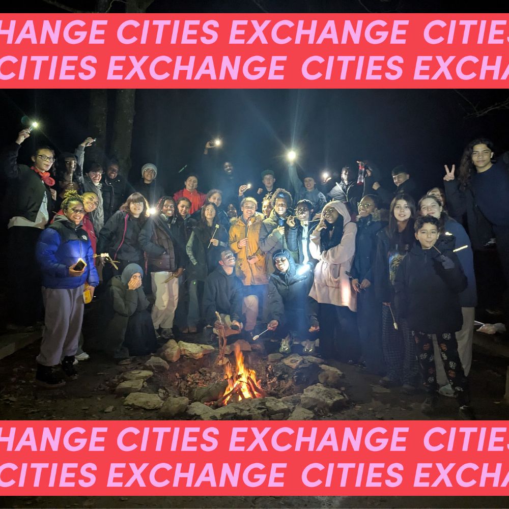 A group of young people stand around a campfire at night, holding flashlights. Within the group there is a wide range of skin tones. A banner reads "CITIES EXCHANGE" in bold pink text.
