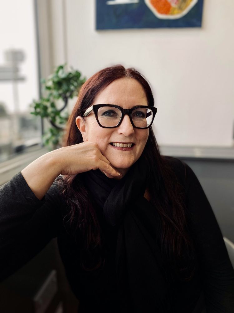 A woman with long burgundy red hair, dark eye makeup and black glasses smiles. Her chin is resting on her hand in a white room. Behind her there is a plant, large window, and blue abstract painting.