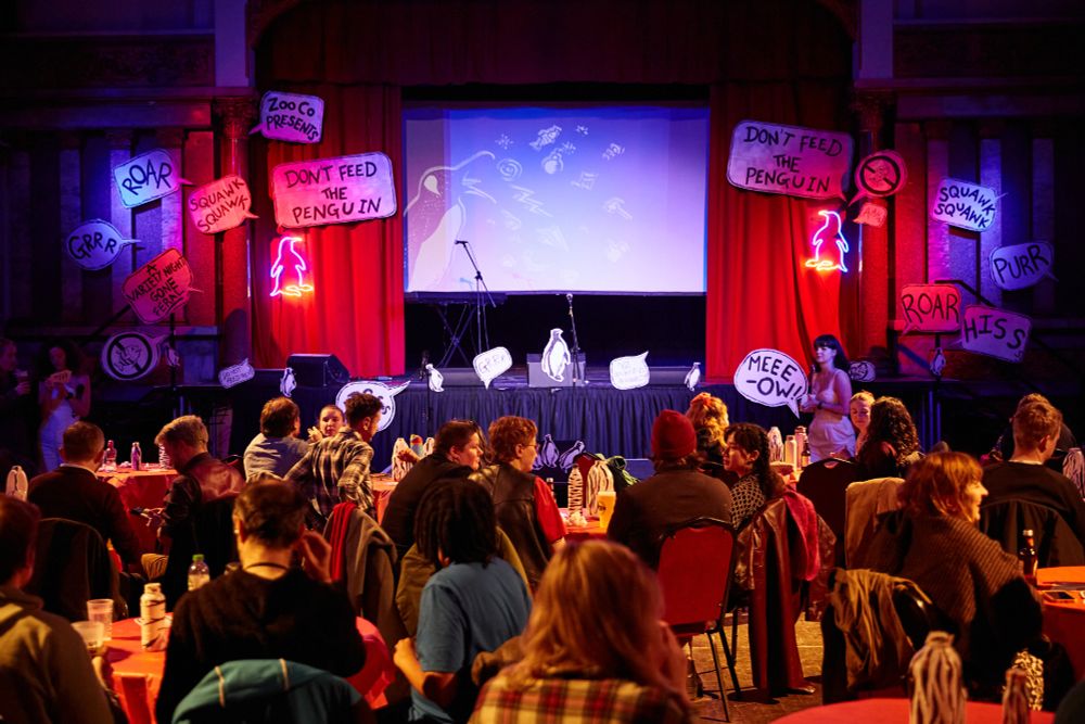 A phot from behind the audience of the stage space. People are chatting sat around round tables with red table cloths. The stage space is lit with red and blue light. Three microphones are in the space and the stage is surrounded by white speech bubbles with animal noises written in. Two neon penguin lights are either side of the stage.