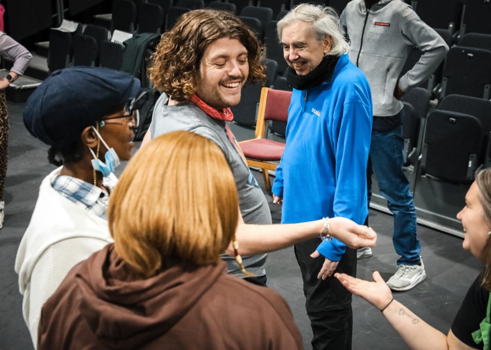 A group of people smiling and interacting infront of theatre seating. A white man with curly hair and a red bandana round his neck grins in the centre of the group.