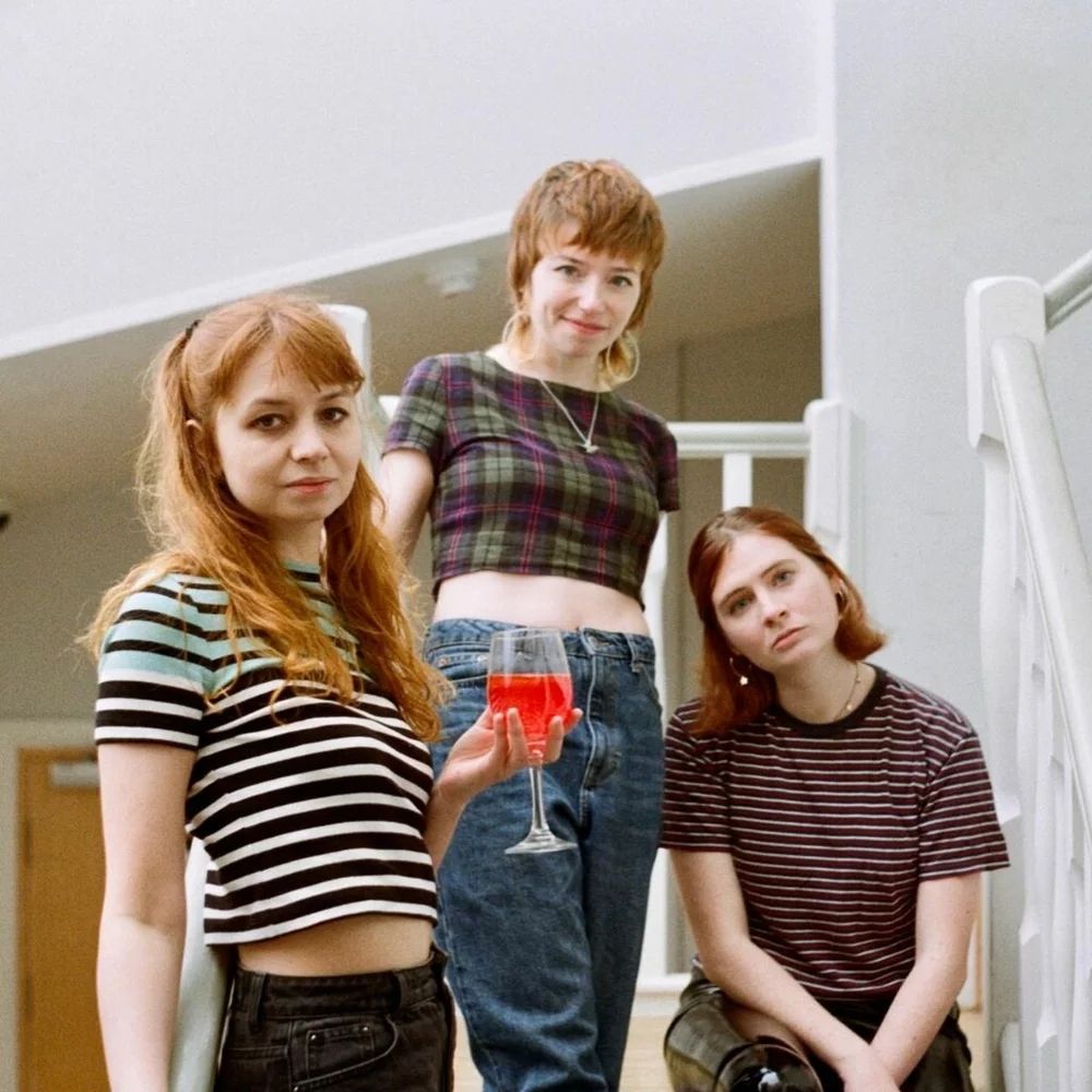 A band photo of trio Cheerbleederz, each wearing different patterned tops, Soph is holding a wine glass of bright red liquid