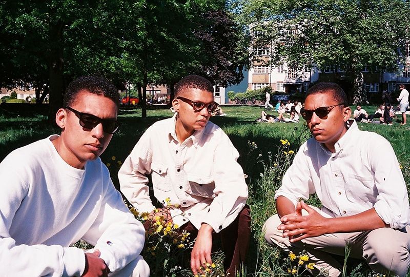 A band photo of Sister Wanzala, all three wear white clothes and sunglasses. They're squatting in a green park on a sunny day