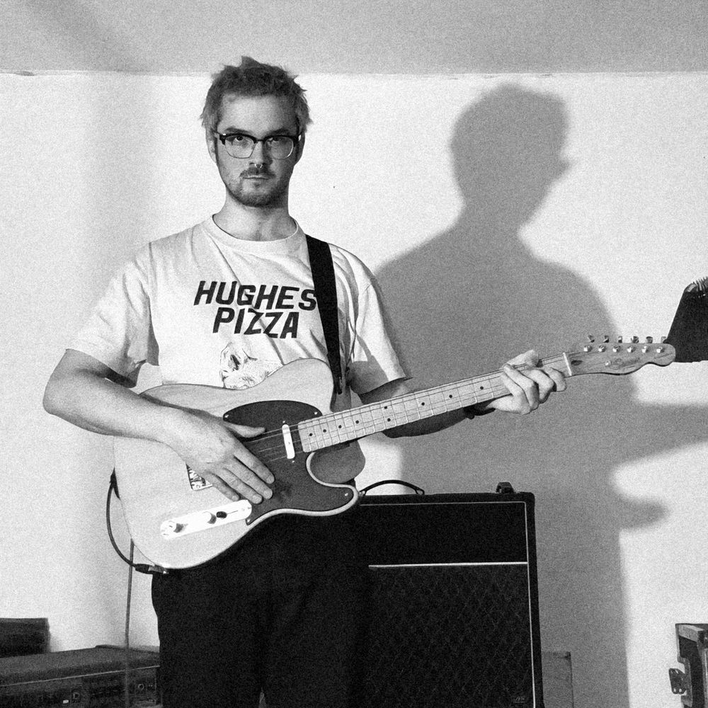 A photo of Jay Cavalier, he's holding a telecaster guitar stood in front of a Vox amplifier and wearing a t-shirt that days "Hughes Pizza". He has glasses on, and short facial hair
