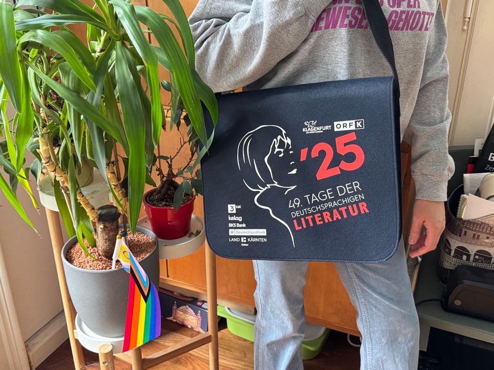 eine person steht nemen einer topfpflanze und trägt die umhängetasche zu den 49. tagen der deutschsprachigen literatur