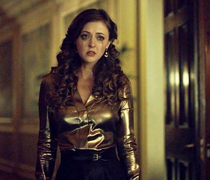 Katharine Isabelle as Margot in Hannibal. She has long curly hair, mascara stained cheeks, one dangly earring and is wearing a shiny gold blouse.